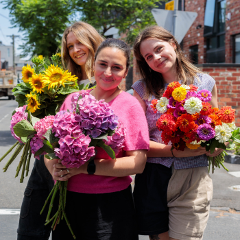 Three women standing on a street, each holding a large bouquet of colourful flowers including sunflowers, hydrangeas and zinnias.